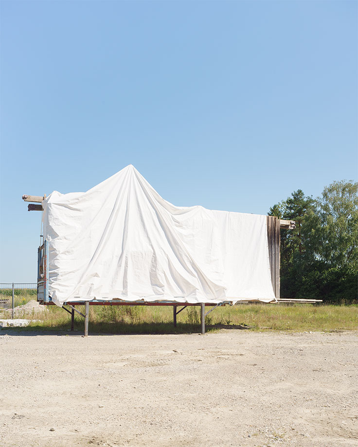 gelagertes Fertigmodulbauteil Holzbau, mit weißer Plane abgedeckt, Foto in der prallen Mittagssonne, Sommer 2025